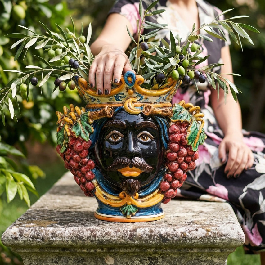 Hand-painted Teste di Moro vase with grape clusters, crown, and olive branches, styled outdoors on stone pedestal
