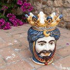 Handcrafted Teste di Moro ceramic vase with crown, blue hair, and red beads, set against a stone wall and flowers.