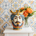 Handmade Teste di Moro ceramic vase with crown, blue hair, and red beads, displayed on white table with orange flowers.