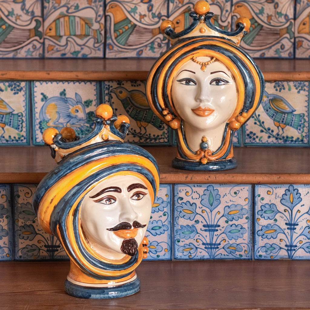 Handmade Teste di Moro ceramic heads with crown, vibrant blue and yellow glaze, displayed on wooden shelf against patterned tiles