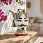Handmade ceramic Testa di Moro vase with colorful jester crown, displayed on rustic wood table with pink flowers.
