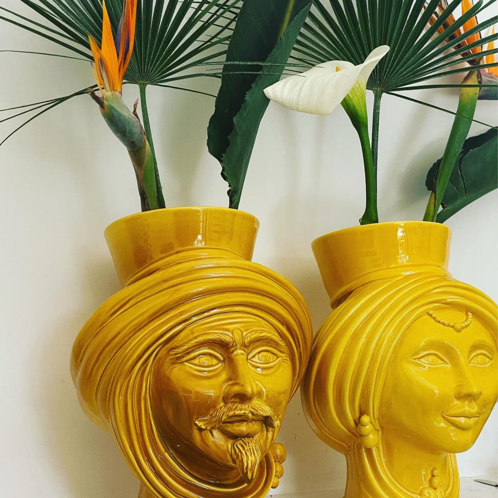 Yellow ceramic Teste di Moro vases with male and female faces, holding tropical flowers, blending tradition and modern design.