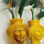 Yellow ceramic Teste di Moro vases with male and female faces, holding tropical flowers, blending tradition and modern design.