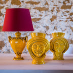 Handmade yellow Teste di Moro vases with ornate faces, paired with a matching lamp, styled on wood against stone wall