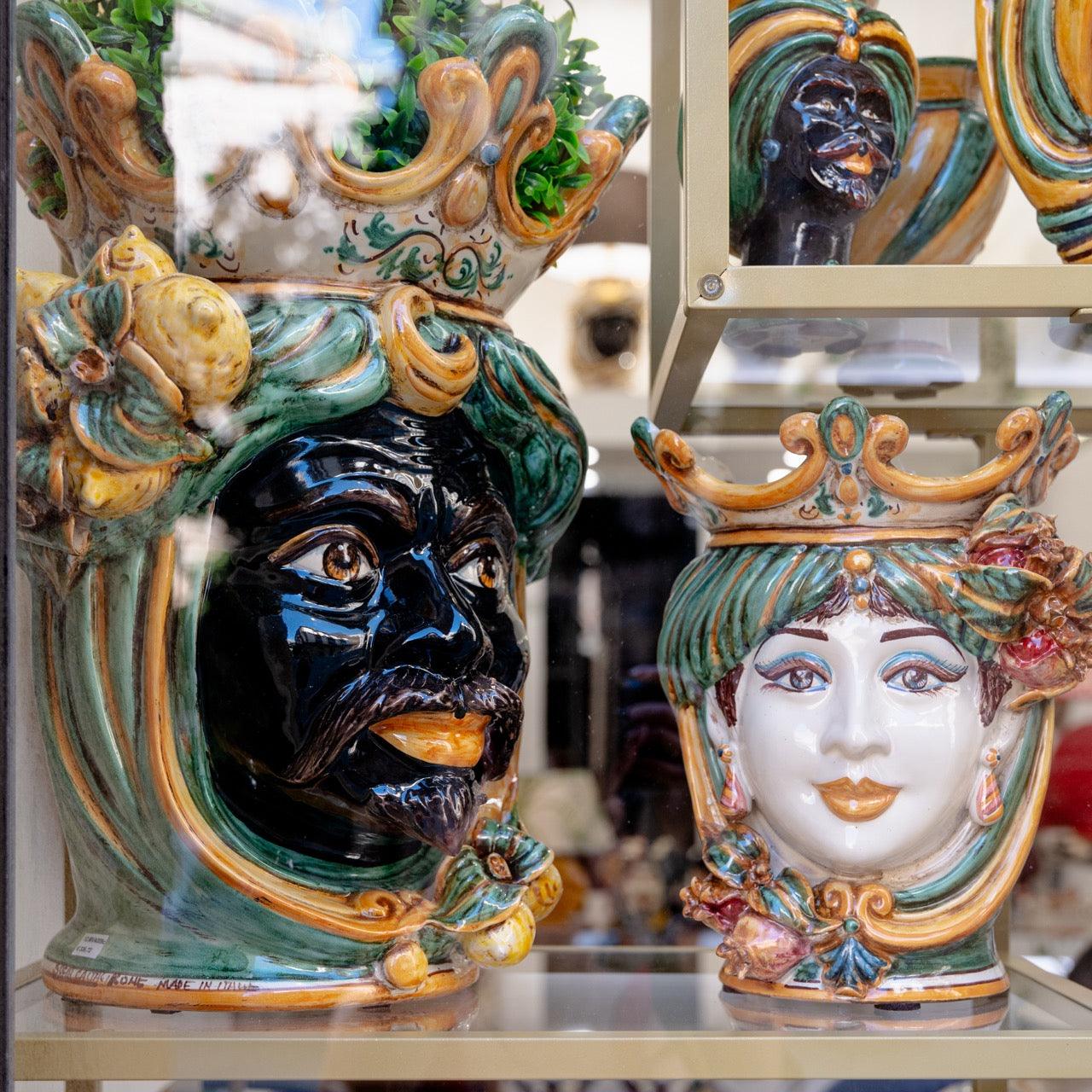 Handmade Teste di Moro ceramic vases with colorful crowns, green foliage, and detailed facial features, displayed in a shop window.