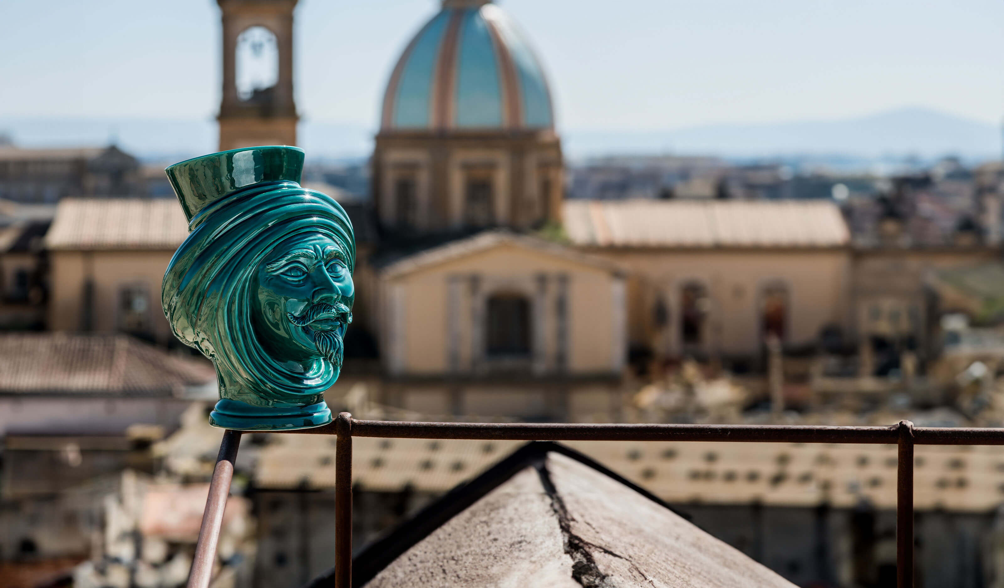 Handmade teal Teste di Moro vase with turban, set against historic cityscape with domed cathedral.